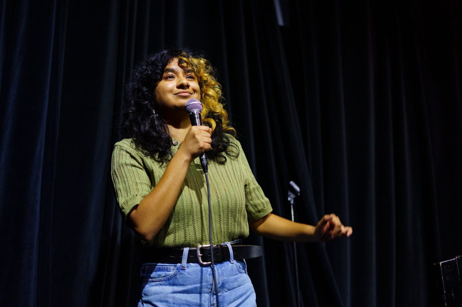 A comedian in a green sweater and jeans holds a microphone on stage.