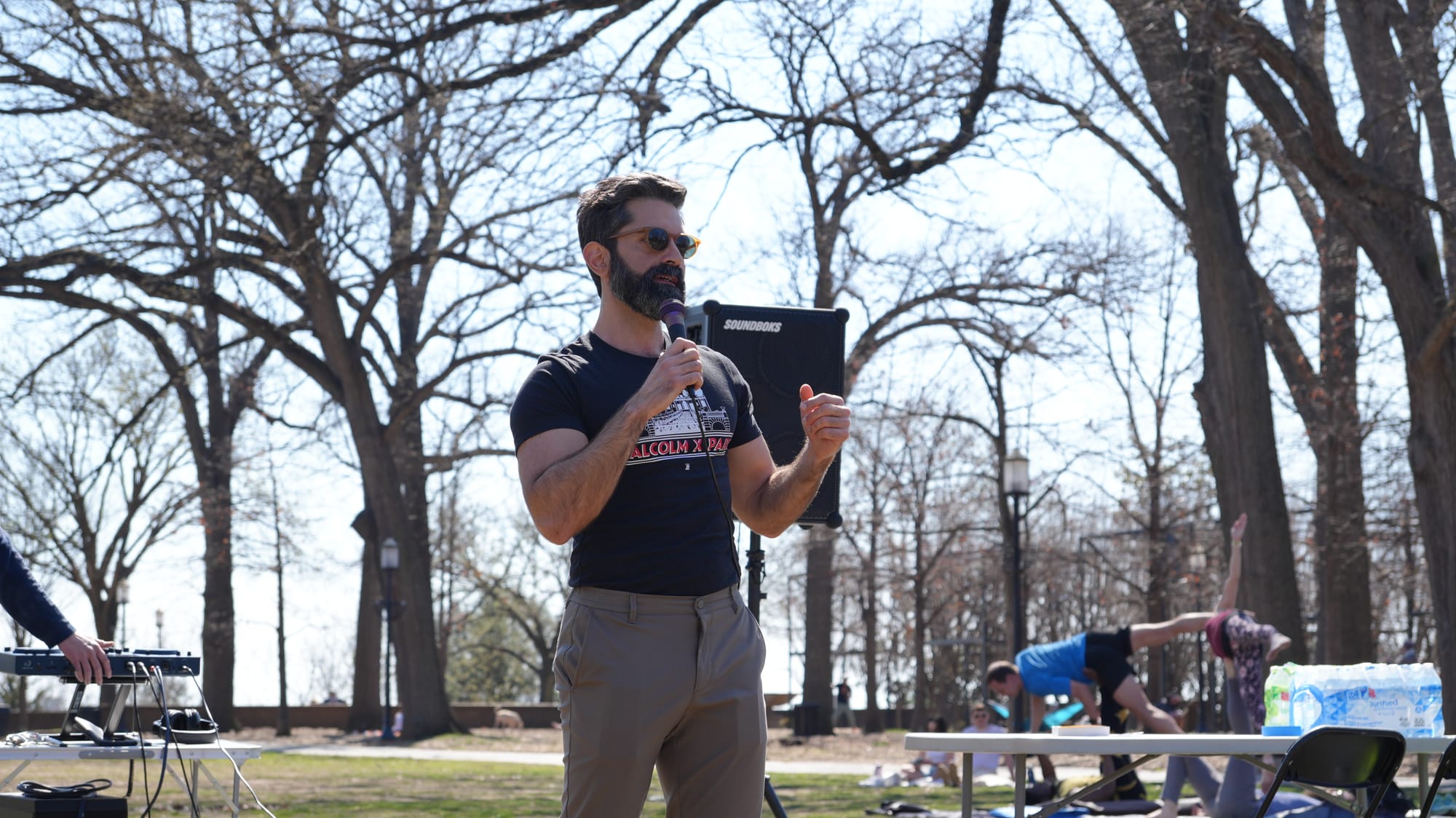 A photo of Miguel Trindade Deramo speaking at a rally at Malcolm X Park. He's wearing sunglasses, a short sleeve shirt that says "Malcolm X Park," and a light grey pants.