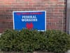 Blue yard sign saying 'This House Supports Federal Workers' against a brick wall with a green bush in front.