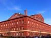 A phot of the The National Building Museum, a large, red brick building with a lot of windows, with blue skies in the background.