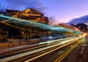 Streaks of lights on a road with parked cars and string lights hanging from a building.