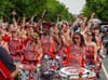 A group of women play drums in matching red, black, and white outfits that read Batalá.