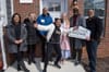 A group of people of different races stand in front of a brick house, holding a sign that says WELCOME HOME.