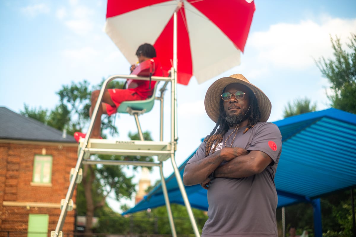 an wearing hat with arms folded standing next to lifeguard stand at pool.