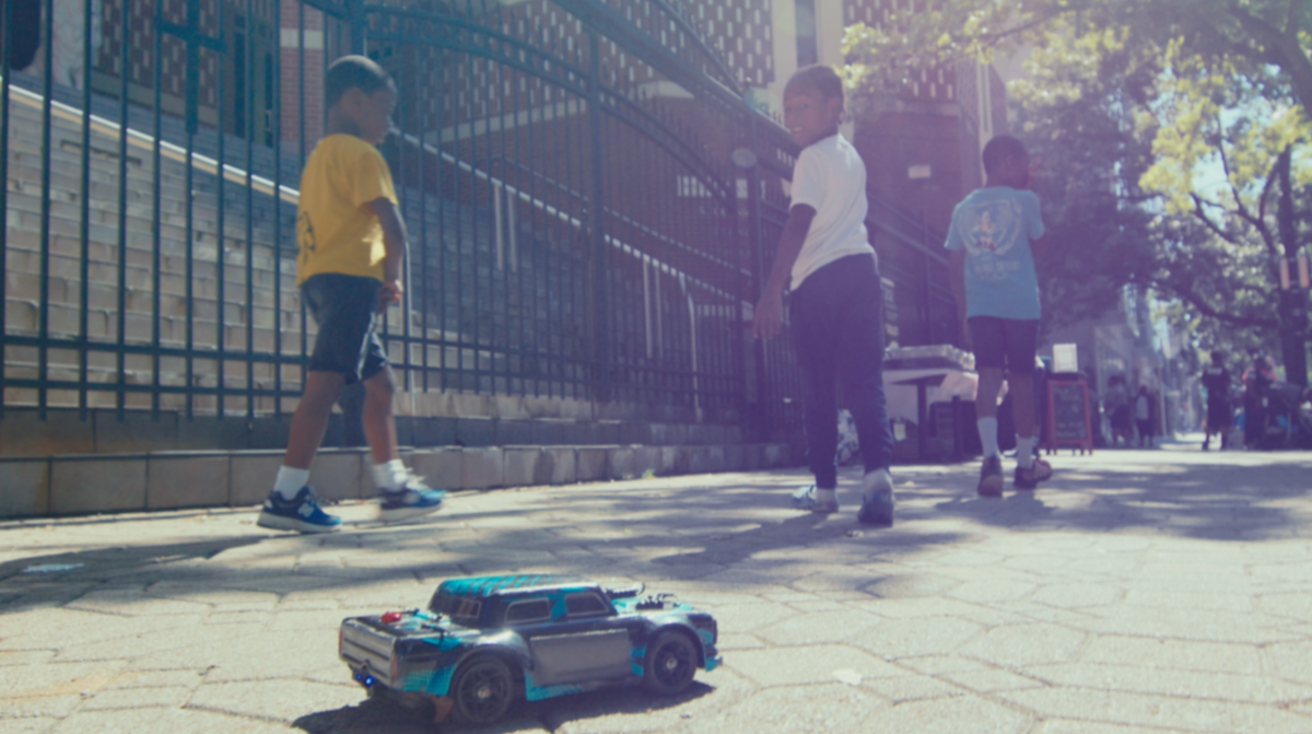 Three kids playing on a street look back at a toy car. 