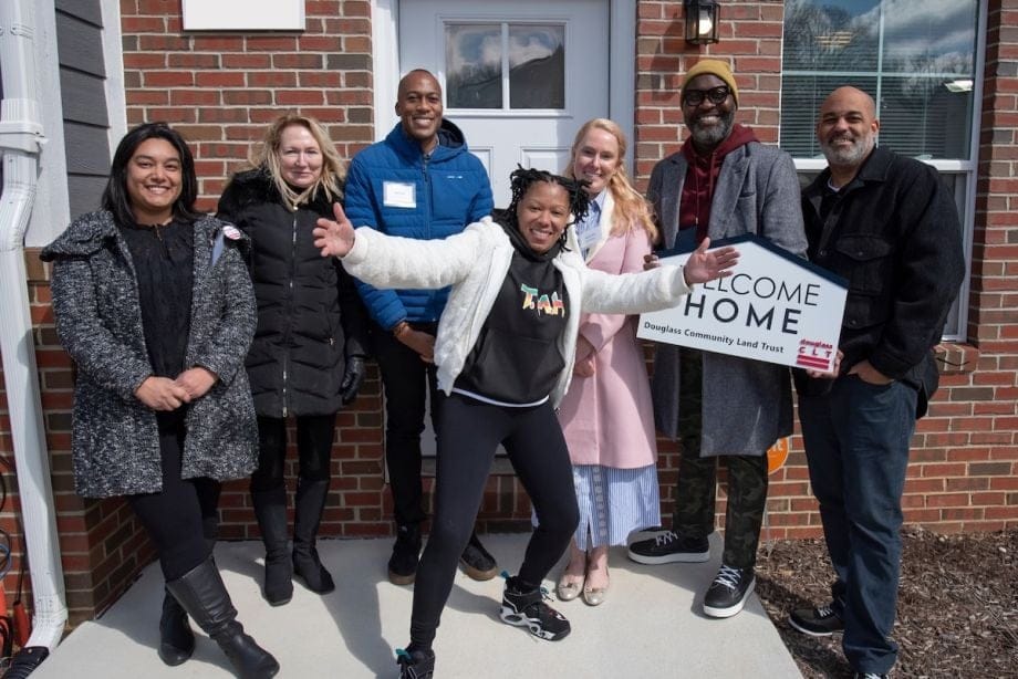 A group of people of different races stand in front of a brick house, holding a sign that says WELCOME HOME.