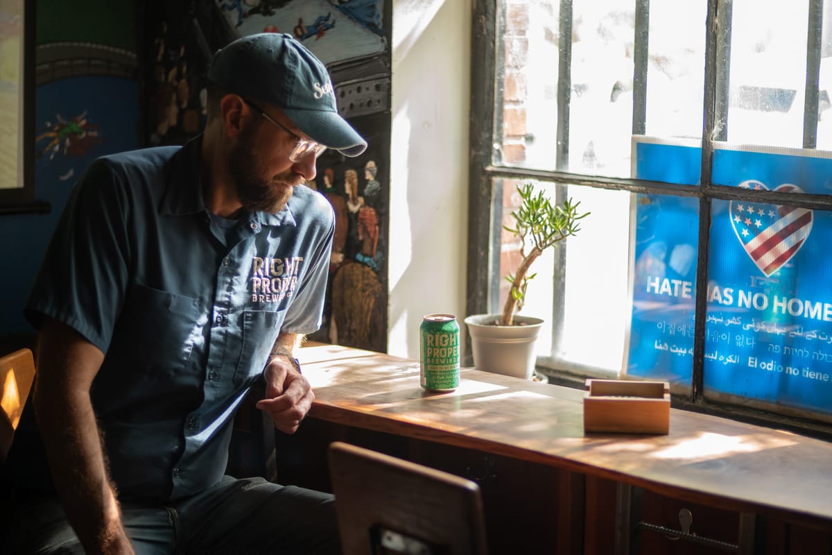A man in a Right Proper Brewer jumpsuit and hat looks down at a can of beer on a table.