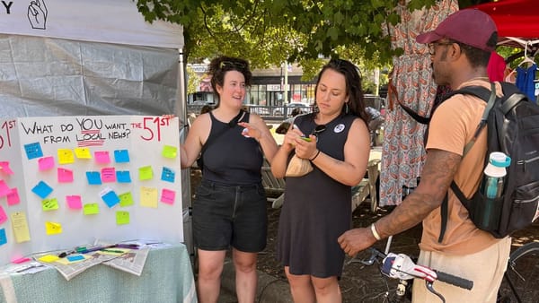 Photo showing two people standing next to a board that reads "Whad to YOU want from local news?", talking to a man.