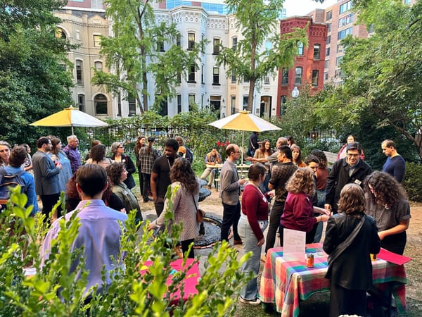 A picture of a couple dozen people gathered in a garden on a sunny day, with colorful row houses in the background.