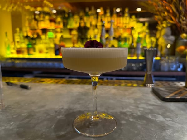 A cocktail glass sits on a bar. The drink is yellow on the base and becomes white and foamy at the top.