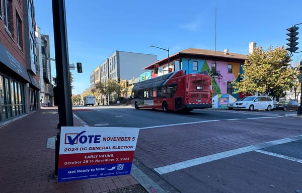 A photograph of a street in Ward 8 with a bus in the background and a sign that says "Vote November 5" in the foreground.