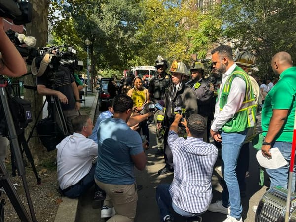 A media availability on the street outside the building. Reporters stand around a group of firefighters.