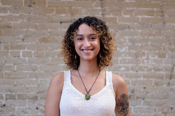 Natalie Delgadillo smiles at the camera in a posed headshot. There is a brick wall in the background.