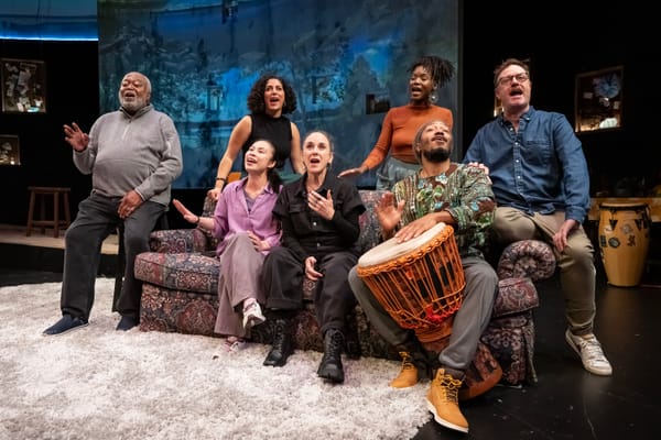 Seven actors sitting on or standing near on a couch while singing together on a theater stage. One is playing a drum.