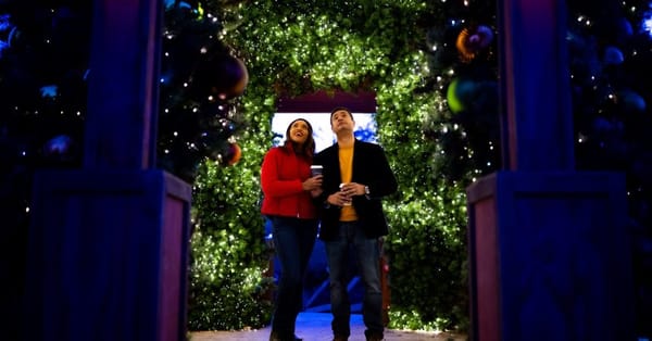 A couple stands in a doorway, smiling upward, with Christmas decorations visible behind and in the foreground.