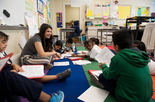 A teacher sits cross-legged on a colorful rug with a circle of students.