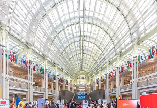 Interior of a glass building with dozens of flags hanging from the ceiling and people visiting booths on the floor.