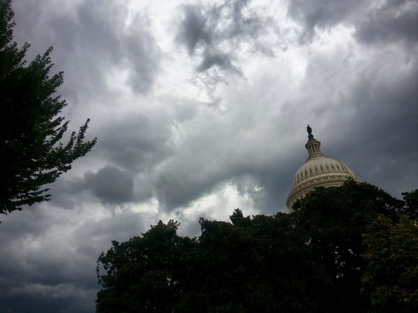 An image of the top of the Capitol dome partially obscured by trees and with a stormy sky in the background.