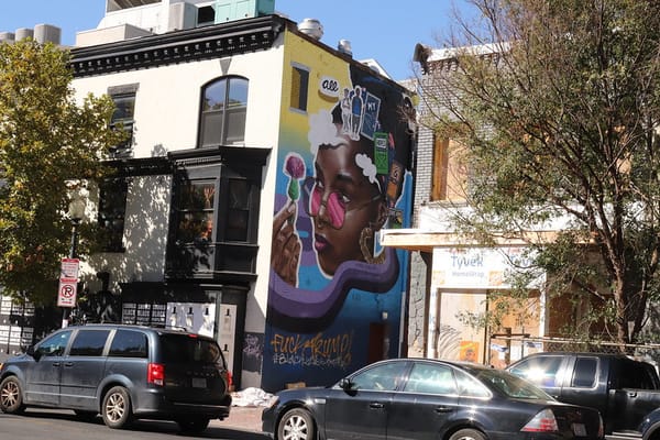 City block with a mural of a Black girl with pink sunglasses painted on the side of a building.