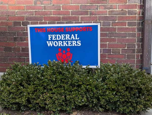 Blue yard sign saying 'This House Supports Federal Workers' against a brick wall with a green bush in front. 