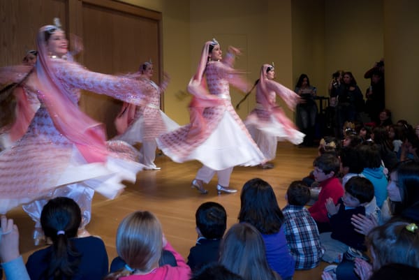 Dancers twirl in pink, white, and glittery dresses in front of children on the floor. 