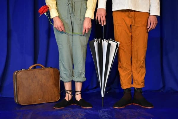 A suitcase and an umbrella stand between the bottom half of two people's bodies. 