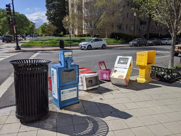 A photo on a D.C. street corner with seven colorful newspaper boxes, five of them are tipped over. 