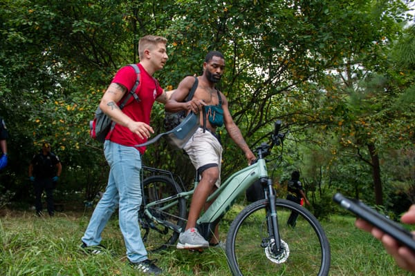 A photo showing two people in conversation in a grove of trees, with multiple officers in the background.