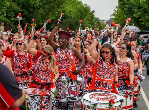 A group of women play drums in matching red, black, and white outfits that read Batalá.