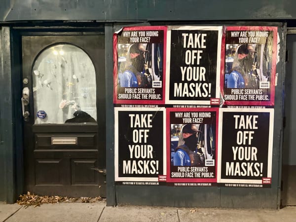 A series of posters on a wall read "Take off your masks!" and "Why are you hiding your face? Public servants should face the public."" 